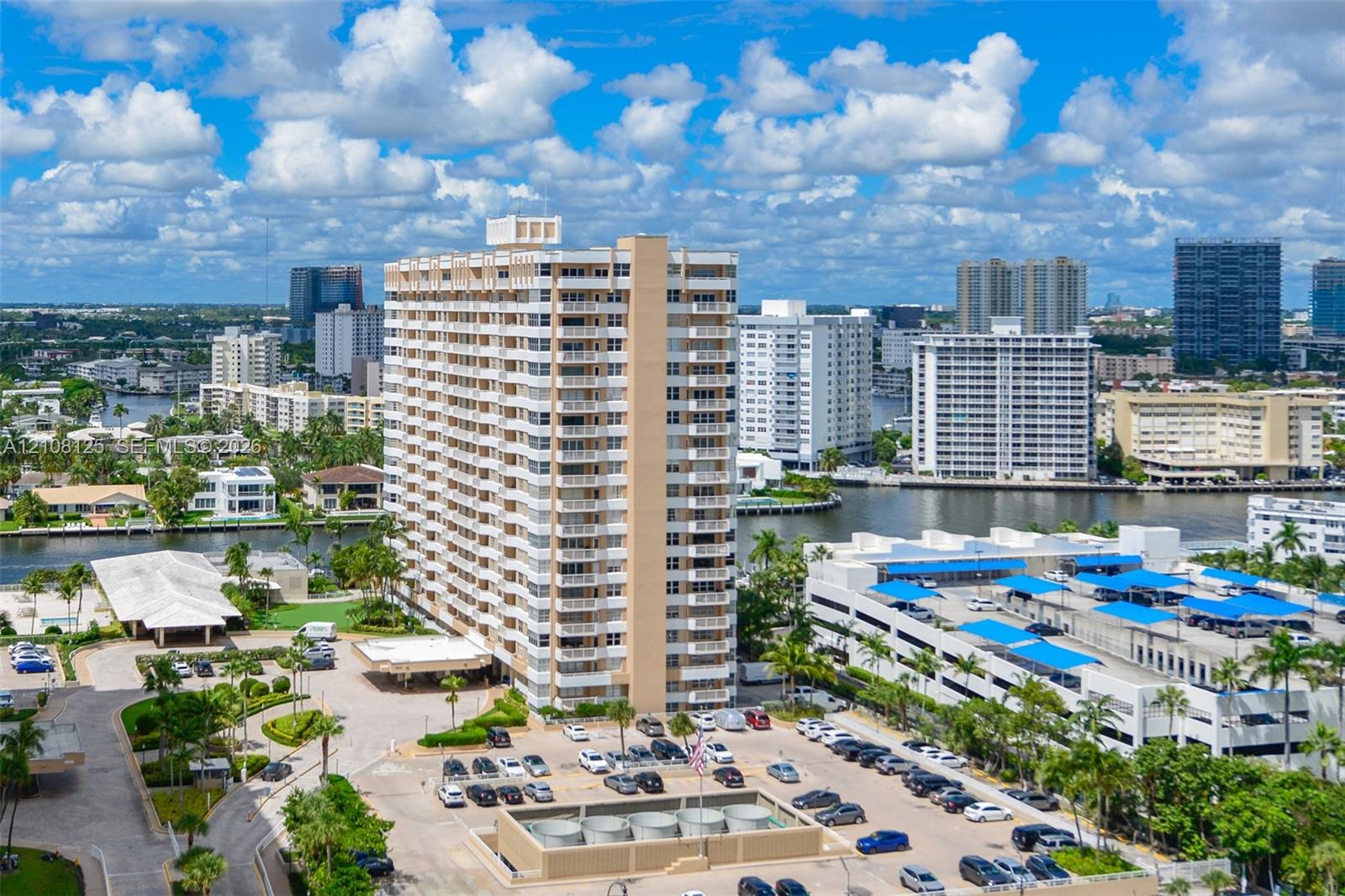 High-rise residential building featuring private balconies, expansive surface parking, a porte-cochre entry, and landscaped grounds set against a waterfront backdrop