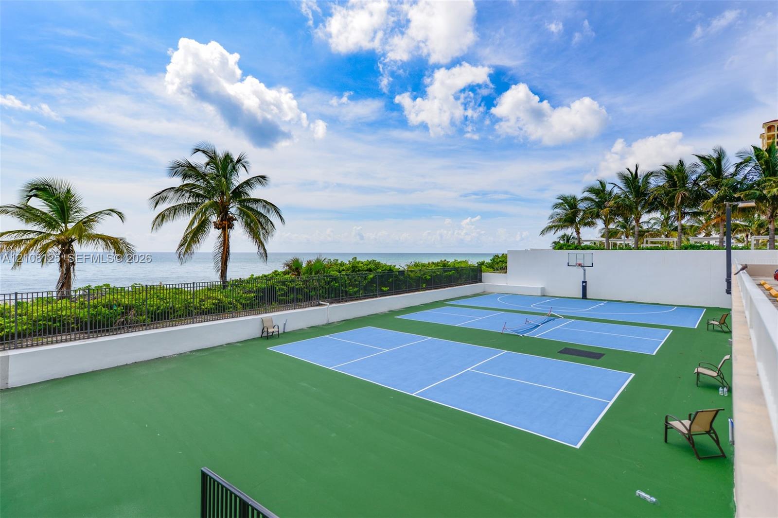 Multi-sport court featuring pickleball and basketball configurations, surrounded by perimeter wall fencing, set against an ocean backdrop with mature palm trees