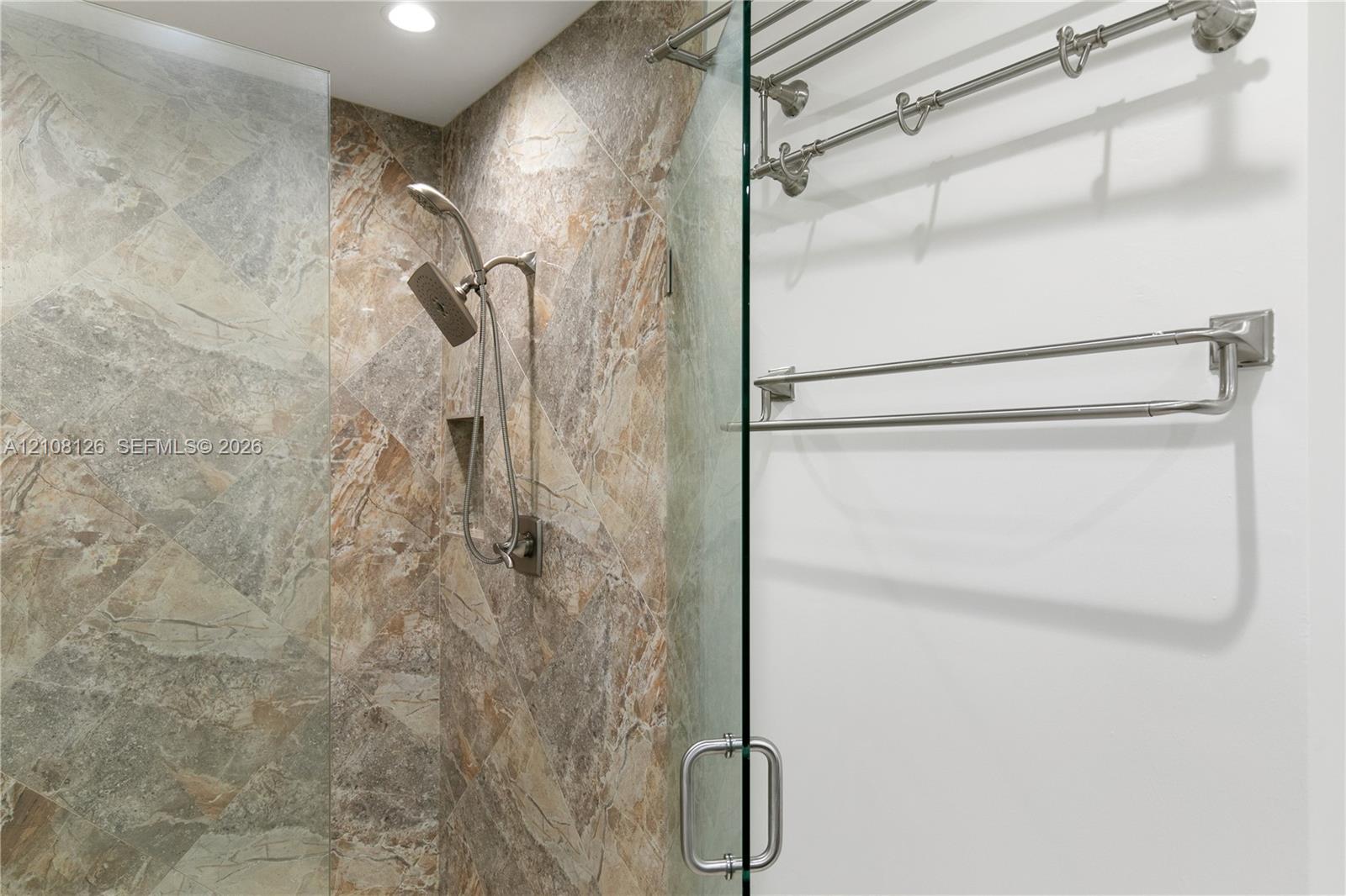 Walk-in shower featuring large-format tile surround, adjustable handheld shower fixture, built-in recessed storage niche, frameless glass door, and brushed nickel hardware