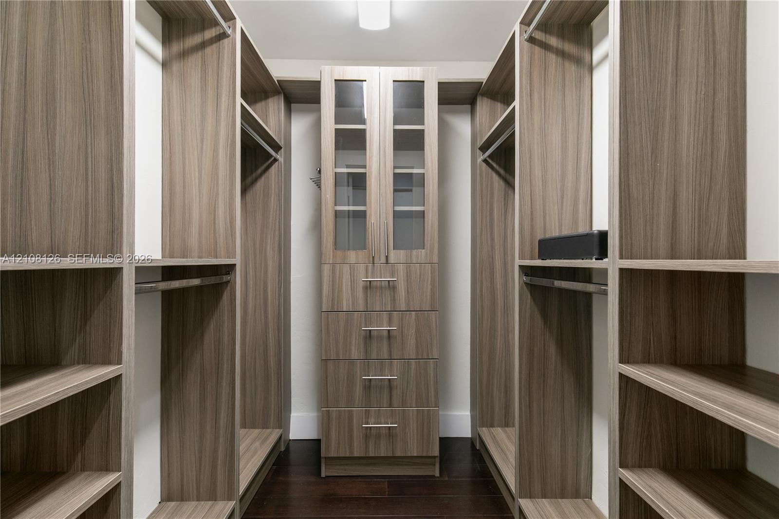 Custom walk-in closet featuring extensive wood-finish shelving, integrated hanging rods, a central stack of four soft-close drawers, and upper cabinetry with glass-panel doors