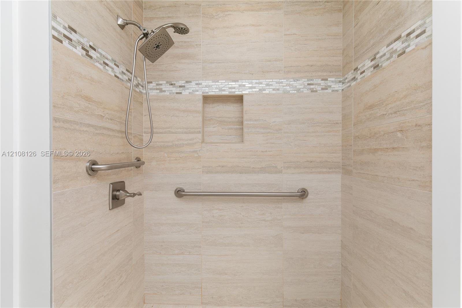 Walk-in shower featuring neutral stone-finish tile surround, integrated mosaic accent banding, built-in recessed niche, handheld shower fixture, and brushed nickel hardware
