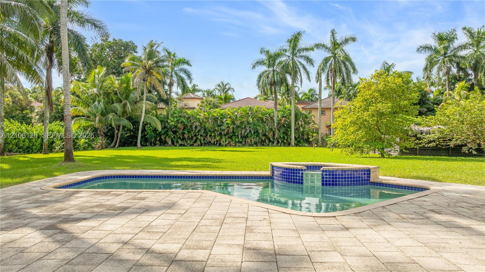 In-ground swimming pool with an attached spillover spa, blue tile detailing, expansive paver patio, and mature palm trees set against a private lawn