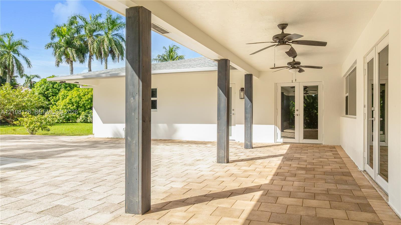 Covered outdoor patio featuring light-toned paver flooring, dark wood-finish structural support columns, dual ceiling fans, and double glass-paned doors