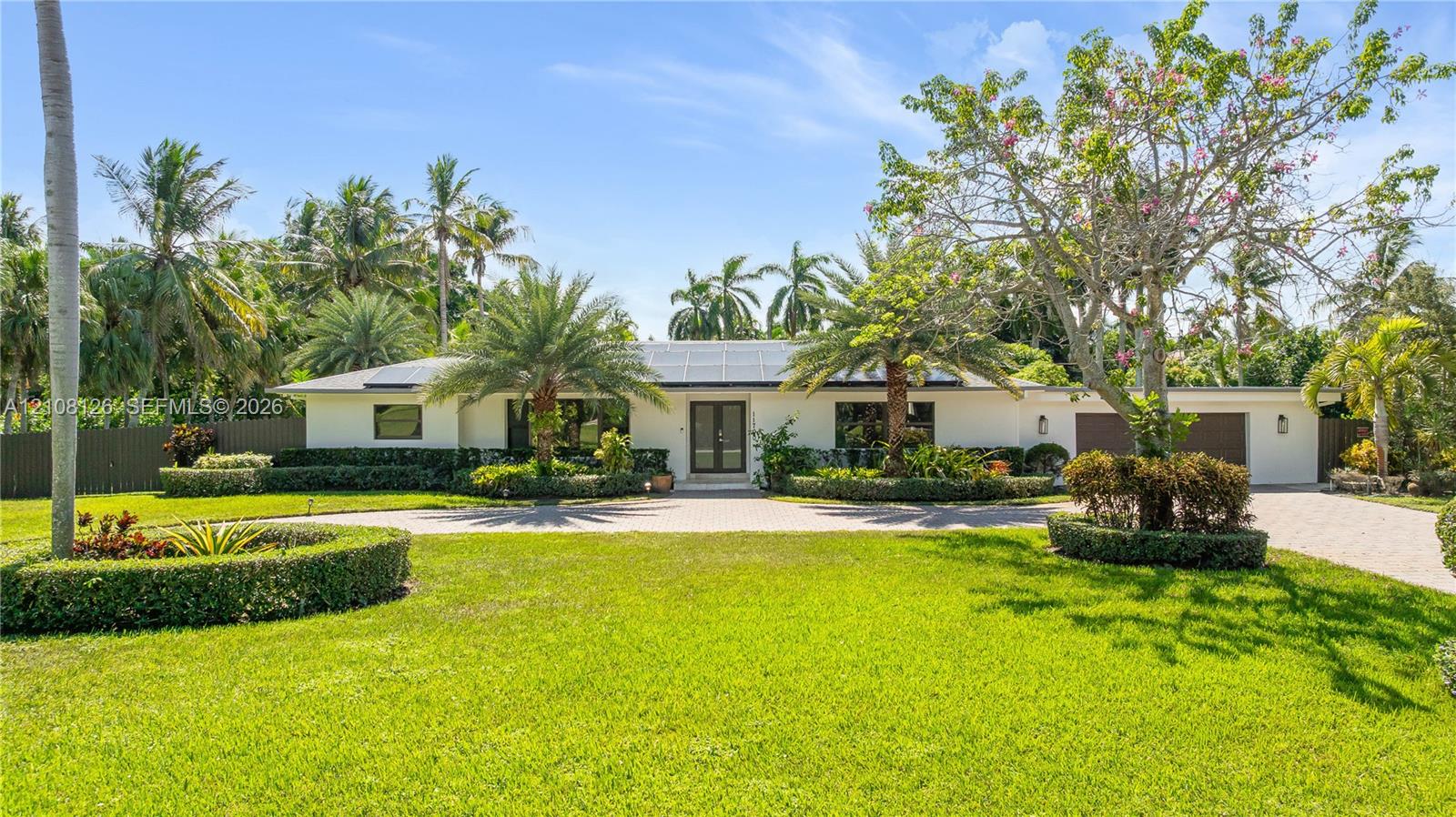 Single-story exterior featuring a light-toned stucco faade, solar panels, paver driveway, manicured landscaping, and mature palm trees