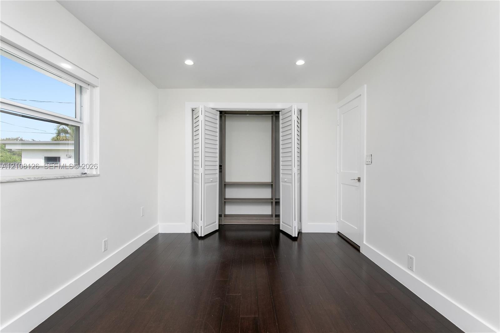 Interior room featuring wood-finish flooring, recessed lighting, white walls, louvered closet doors with built-in shelving, and a dual-pane window