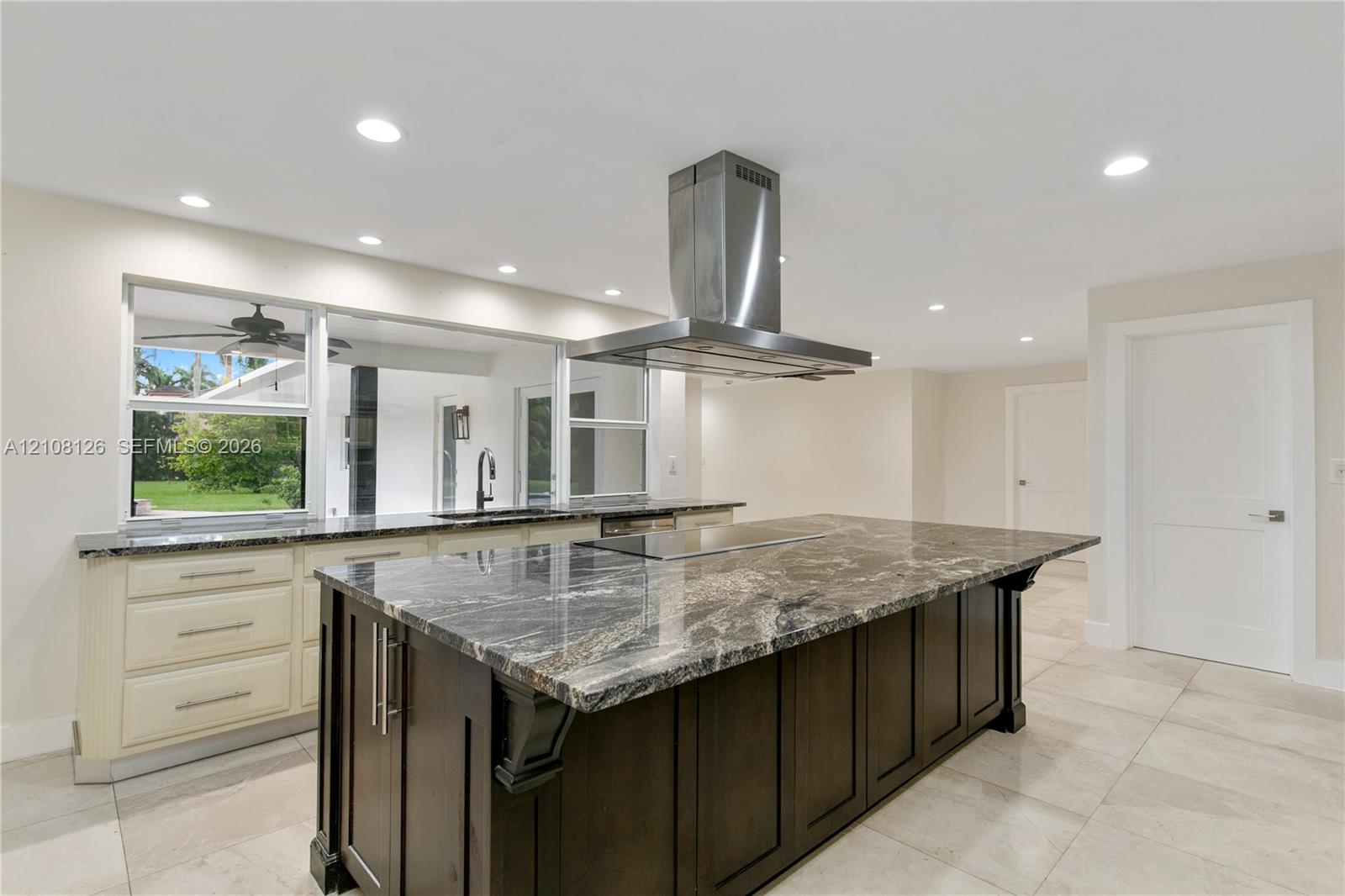 Kitchen featuring a large central island with dark cabinetry and stone-finish countertop, stainless steel ceiling-mounted range hood, light-colored perimeter cabinetry, and large-format tile flooring