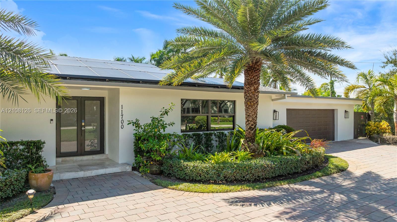 Single-story exterior featuring a light-toned stucco facade, rooftop solar panels, a paved brick driveway, a double-entry glass door, and integrated outdoor wall lighting