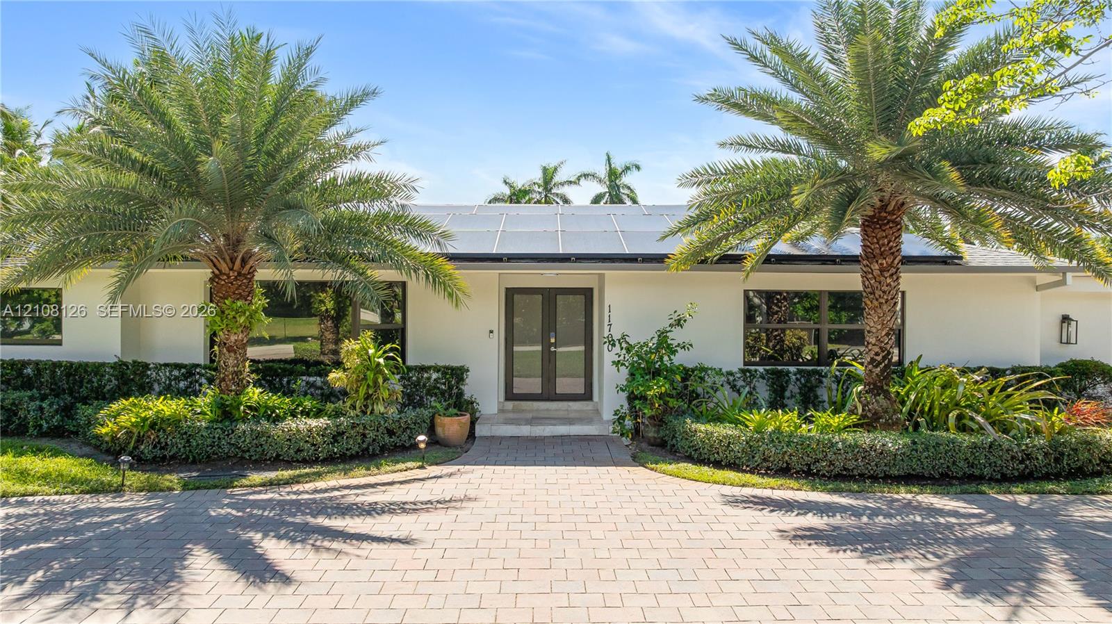 Exterior facade featuring a paved stone driveway, double-door entrance, integrated solar panels, and mature palm trees