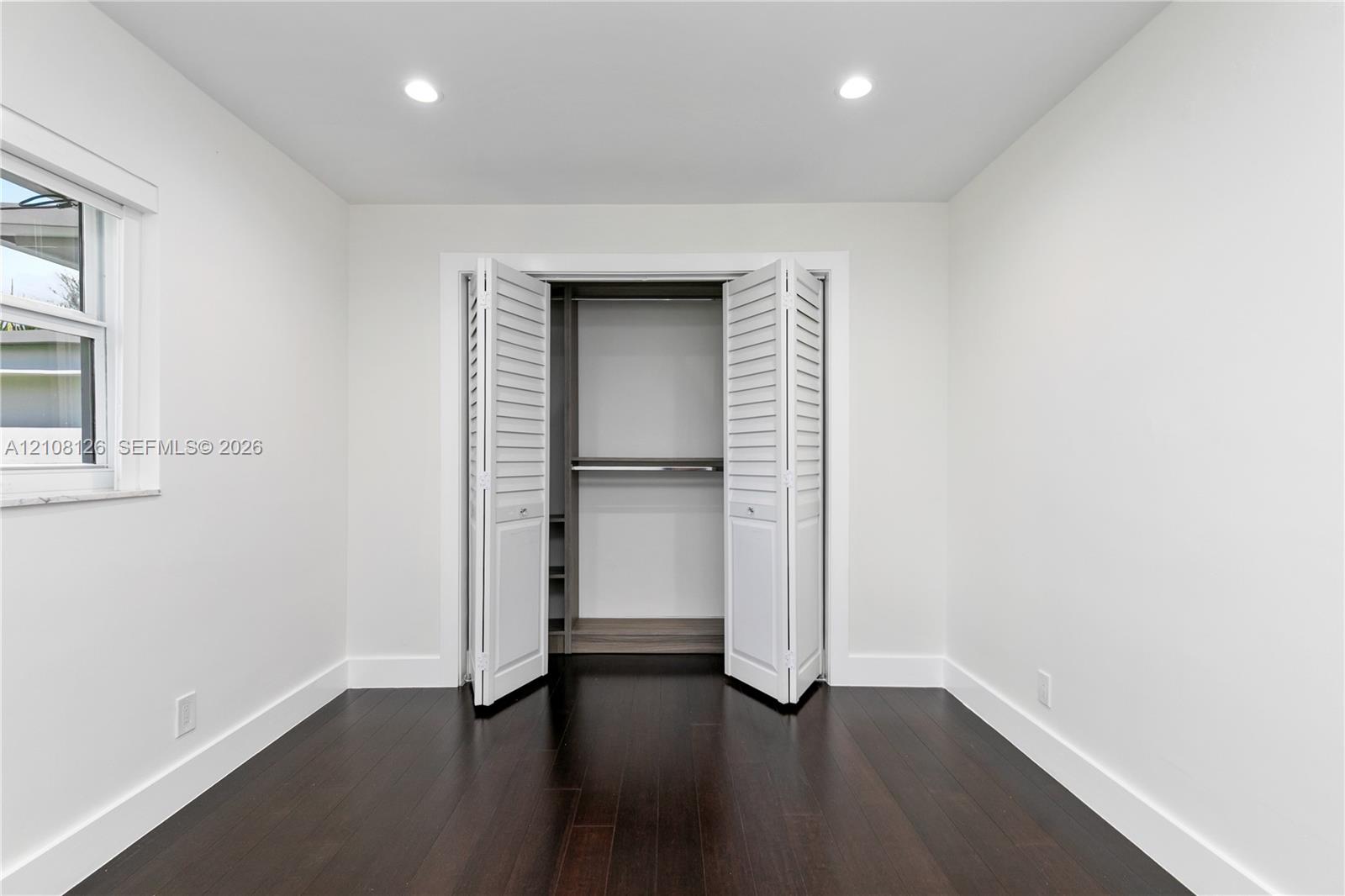 Neutral interior featuring dark wood-finish flooring, recessed lighting, and a closet with white louvered bi-fold doors and built-in shelving
