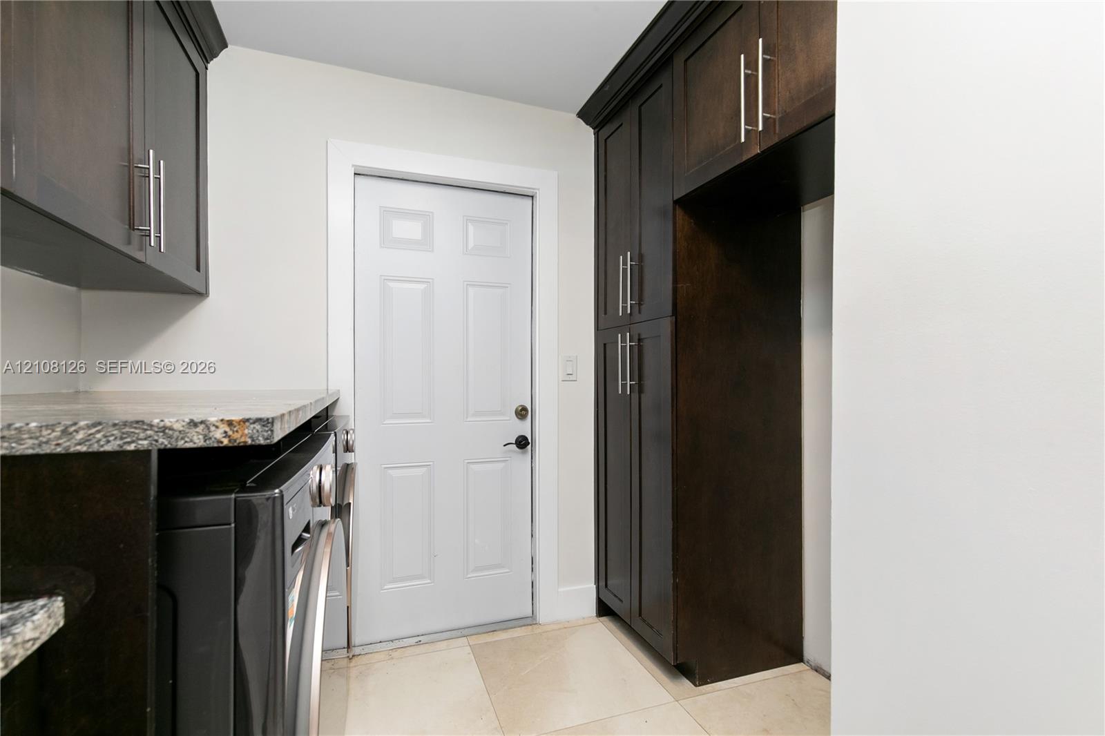Dark-toned cabinetry featuring stainless steel hardware, stone-finish countertops, tile flooring, and a paneled exterior door