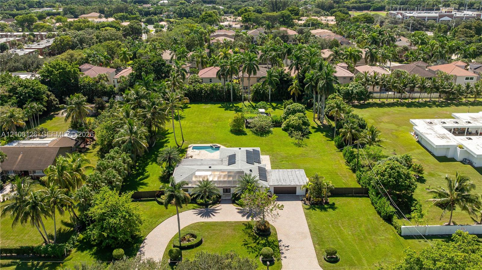 Aerial view of a sprawling property featuring a circular paved driveway, a multi-car garage, solar roof panels, a screened-in swimming pool, and expansive manicured grounds
