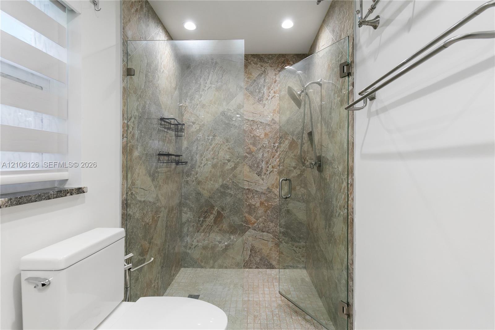 Walk-in shower featuring natural-stone tile surround, frameless glass enclosure, handheld shower fixture, and recessed lighting