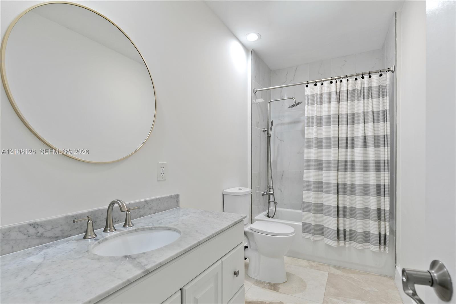 Bathroom featuring a vanity with natural stone countertop, undermount sink, circular wall mirror, and a tub-shower combination with floor-to-ceiling tile surround