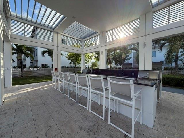 Covered outdoor kitchen featuring stone tile flooring, a built-in counter with a dark stone surface, stainless steel grills, and a slatted pergola roof structure
