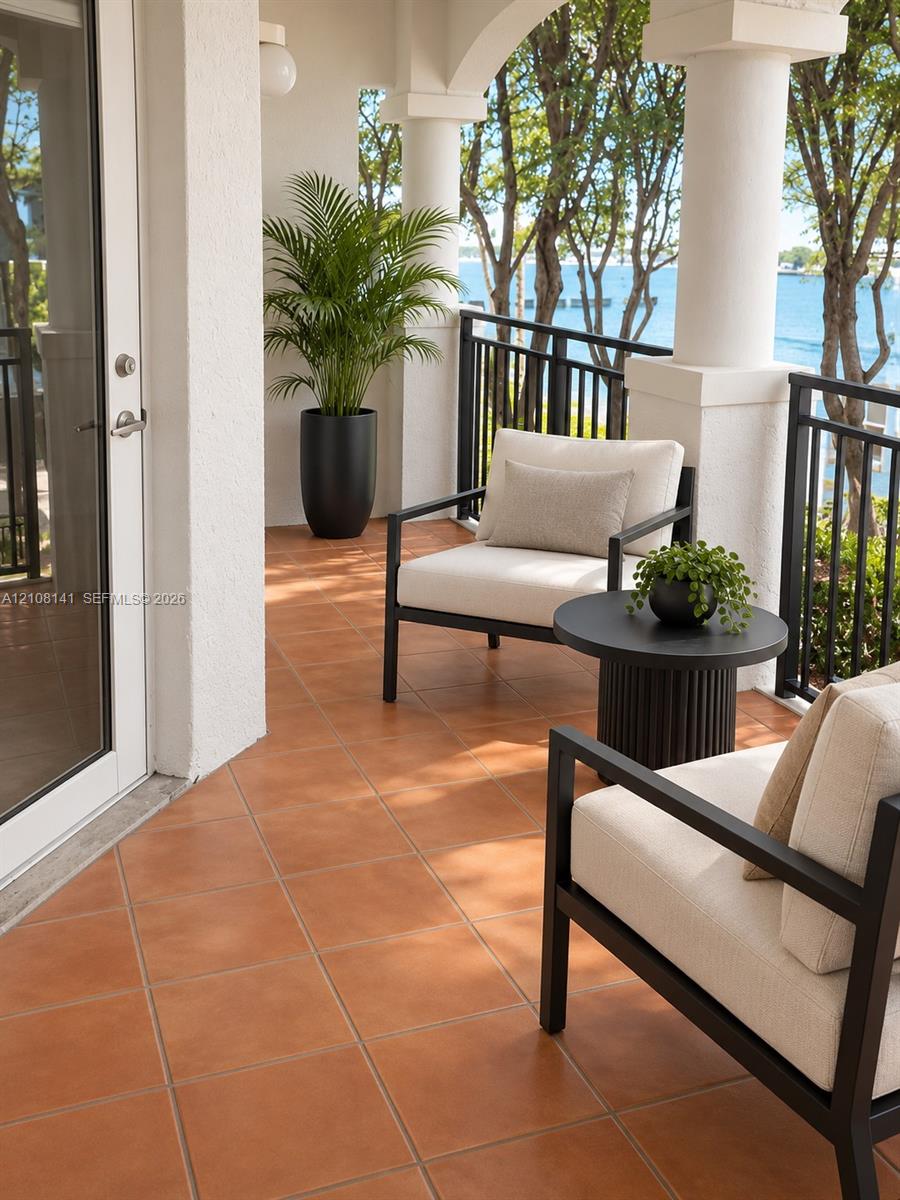 Covered balcony featuring terracotta-style tile flooring, decorative structural columns, and black metal railings with waterfront scenery