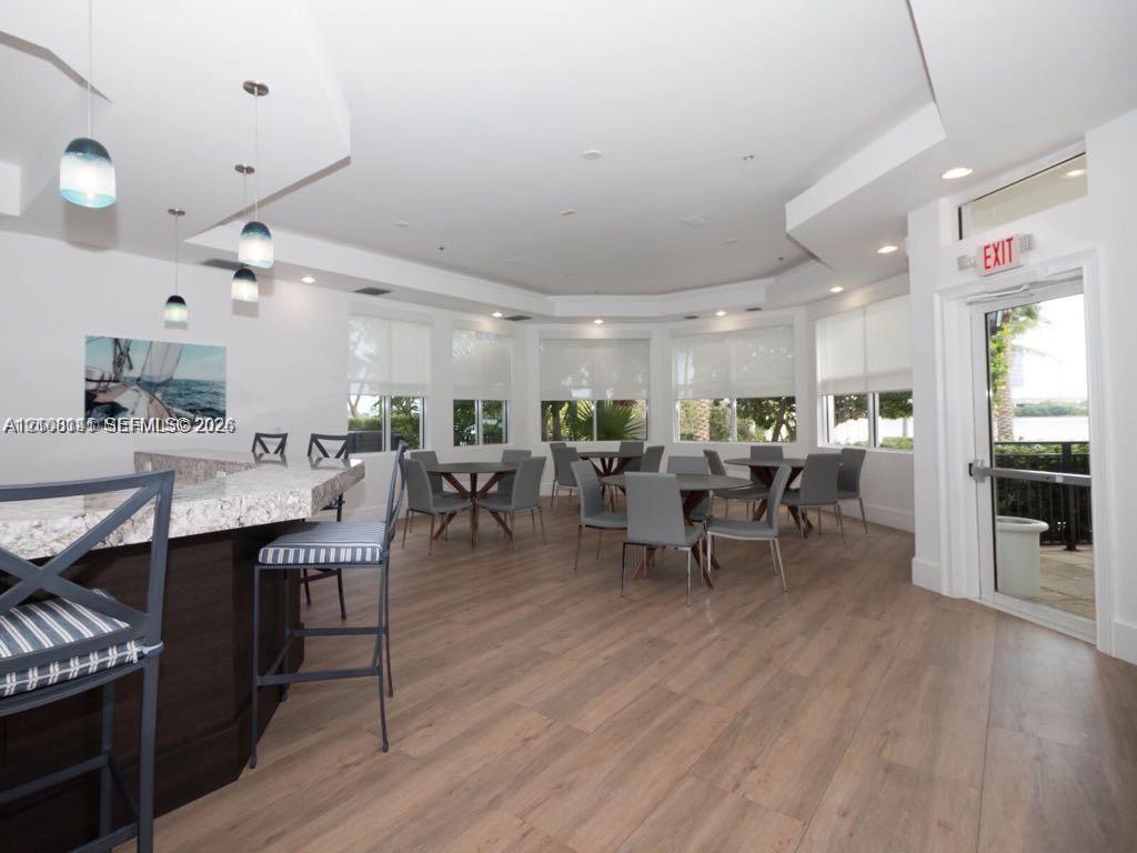 Multipurpose room featuring wood-finish flooring, a built-in bar with stone countertop, multi-tier recessed ceiling, and floor-to-ceiling windows with roller shades