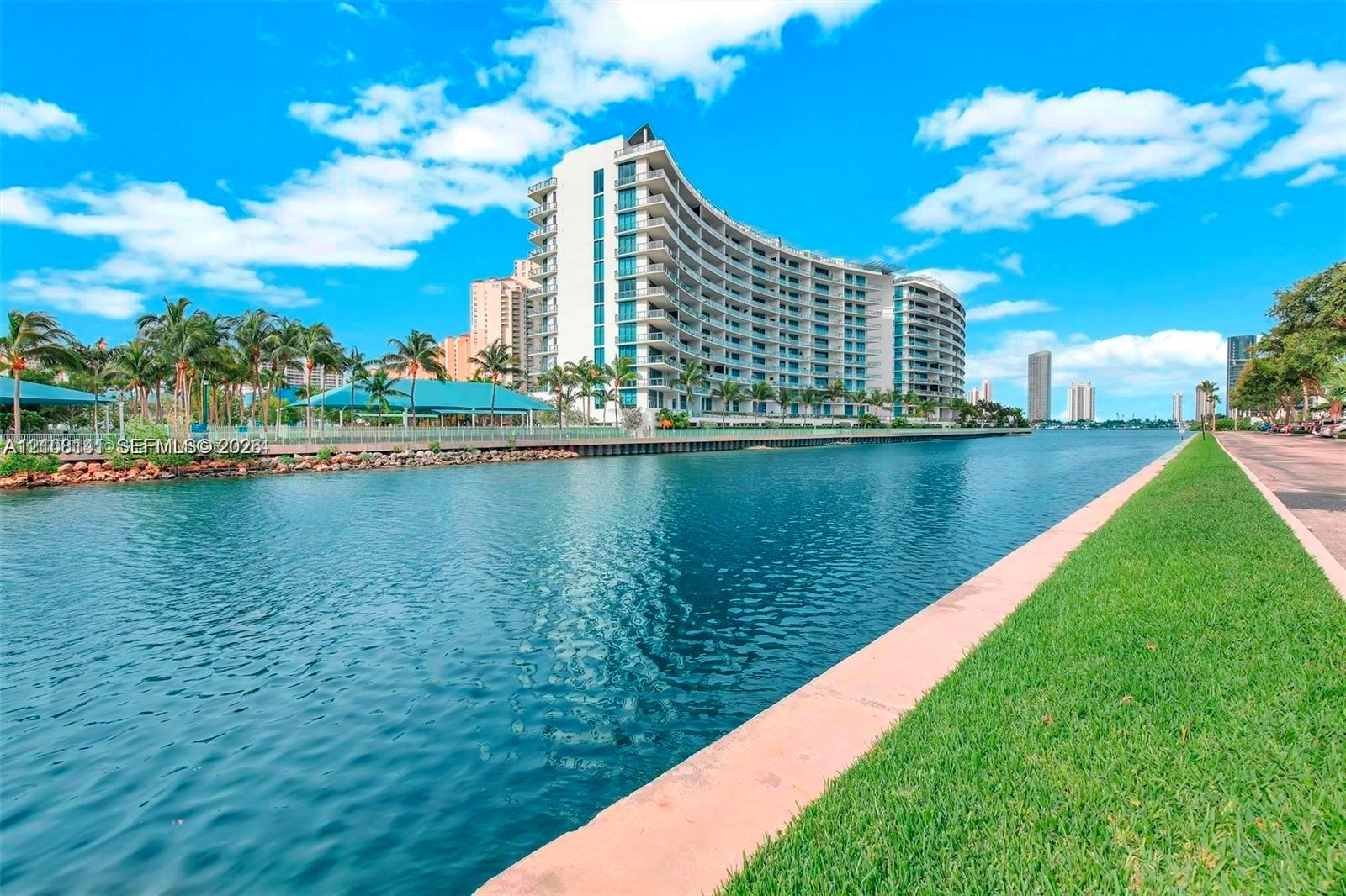 Curved mid-rise architectural facade with expansive balconies, situated on a waterfront lot featuring stone bulkhead, manicured grass landscaping, and established palm trees
