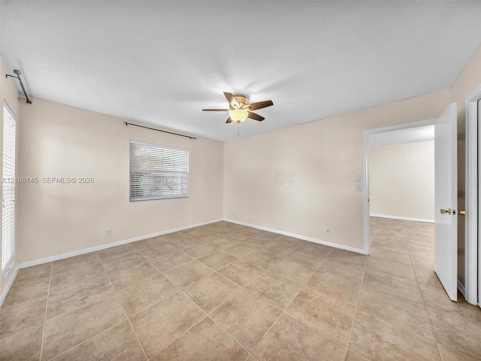 Neutral-toned interior space featuring large-format stone-style tile flooring, dual windows with horizontal blinds, and a central ceiling fan with integrated light fixture