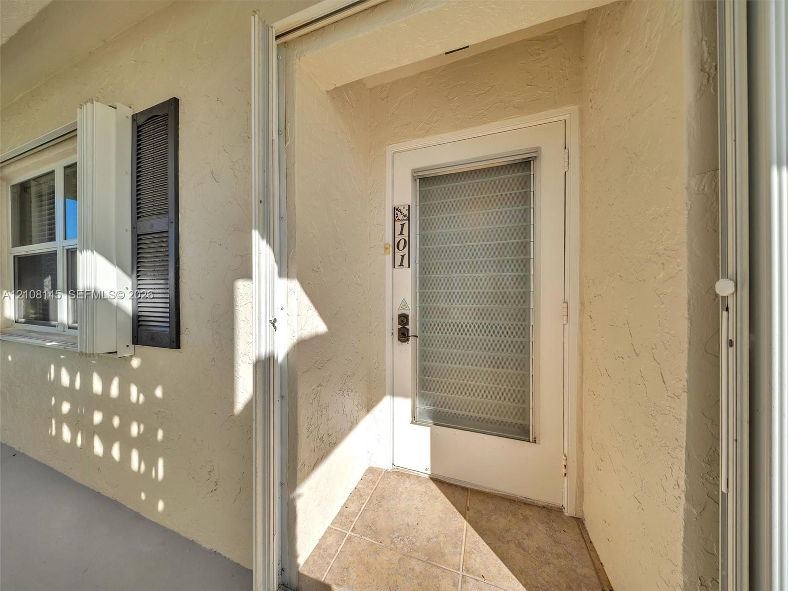 Exterior entry featuring stucco finish, louvered shutters, tile flooring, and a glass-paneled storm door