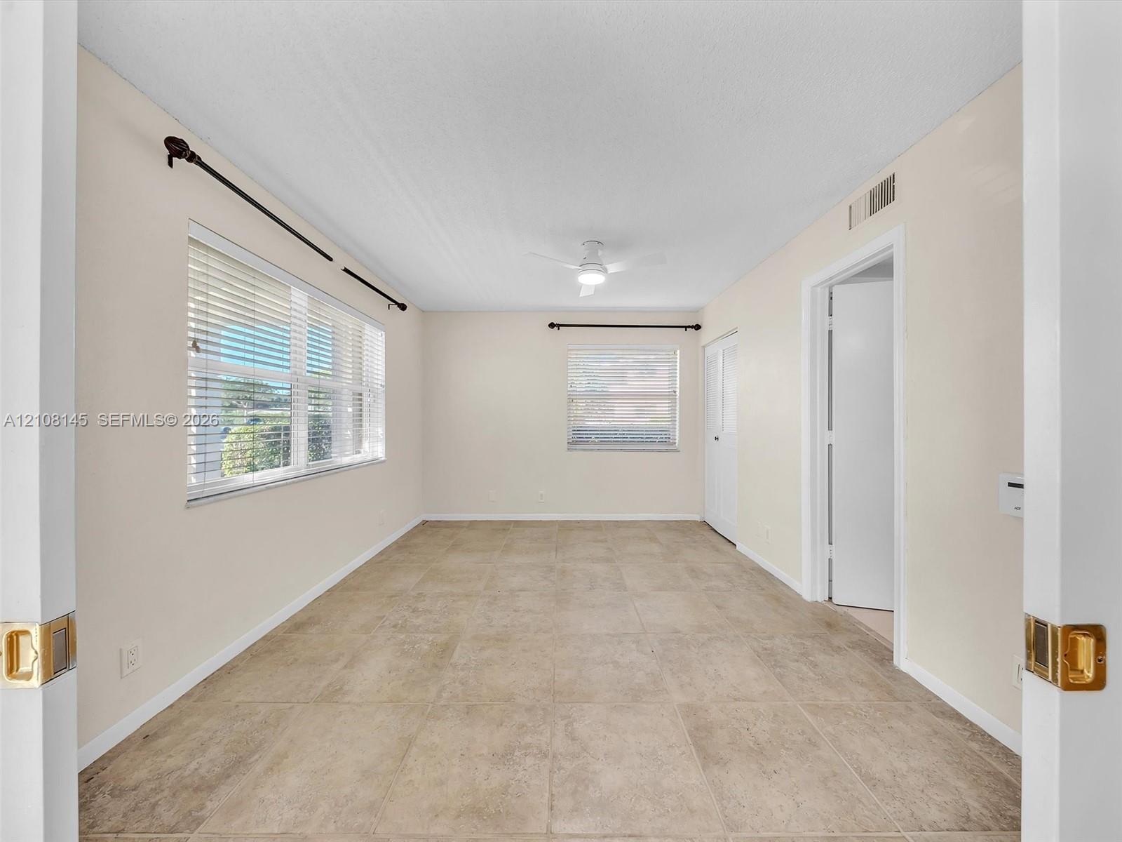 Neutral-toned room featuring large-format tile flooring, dual windows with horizontal blinds, a central ceiling fan with integrated lighting, and white interior doors