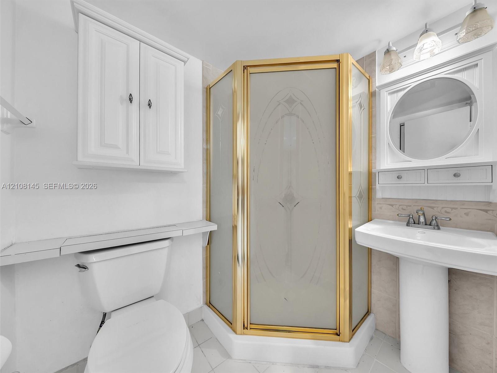 Corner shower with brass-finish frame and etched glass doors, white pedestal sink with dual-handle faucet, circular wall-mounted mirror, white shaker-style wall cabinetry, and tile flooring