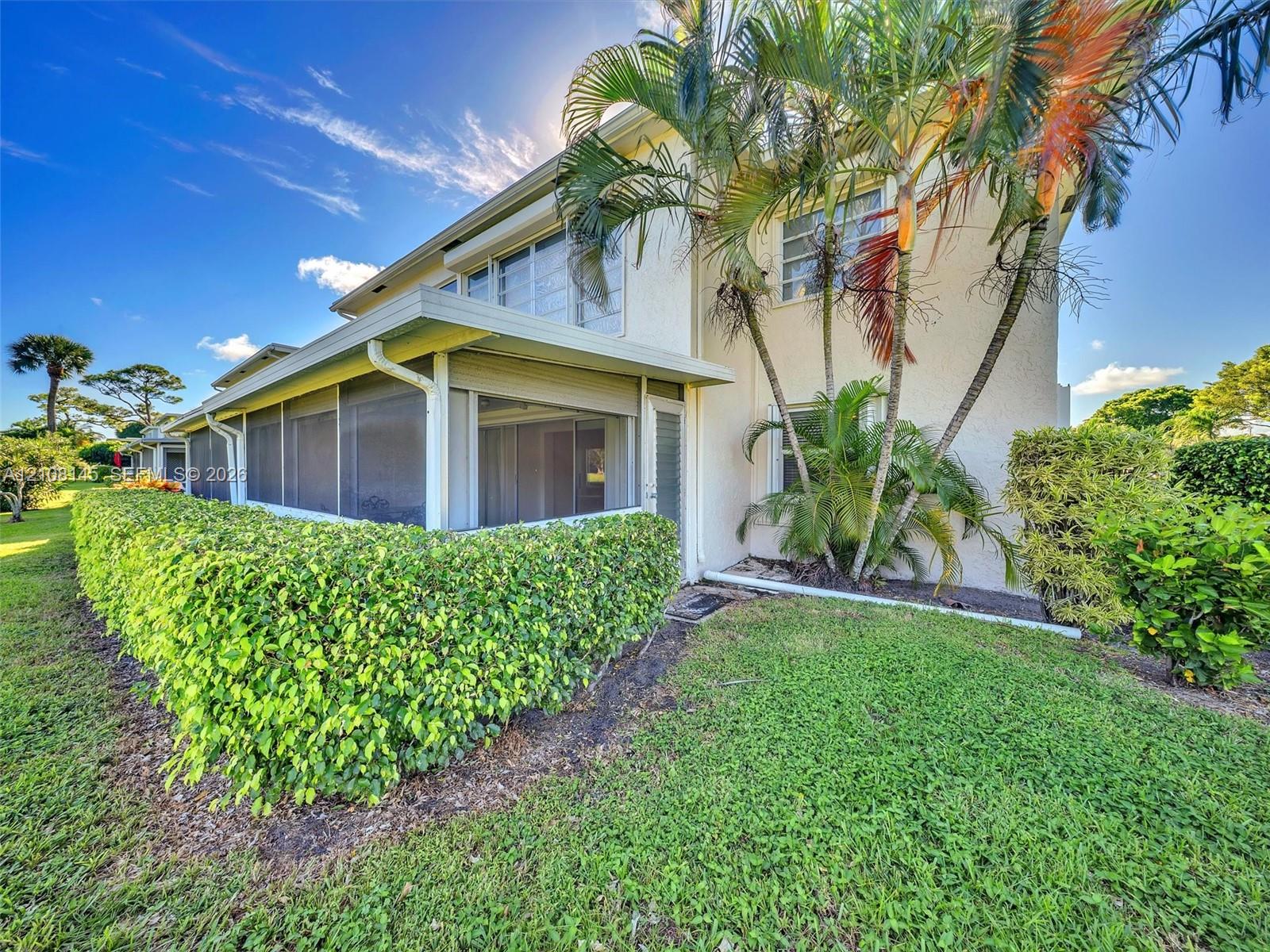 Stucco exterior featuring a screened-in ground level porch, mature palm trees, manicured hedges, and a grass lawn