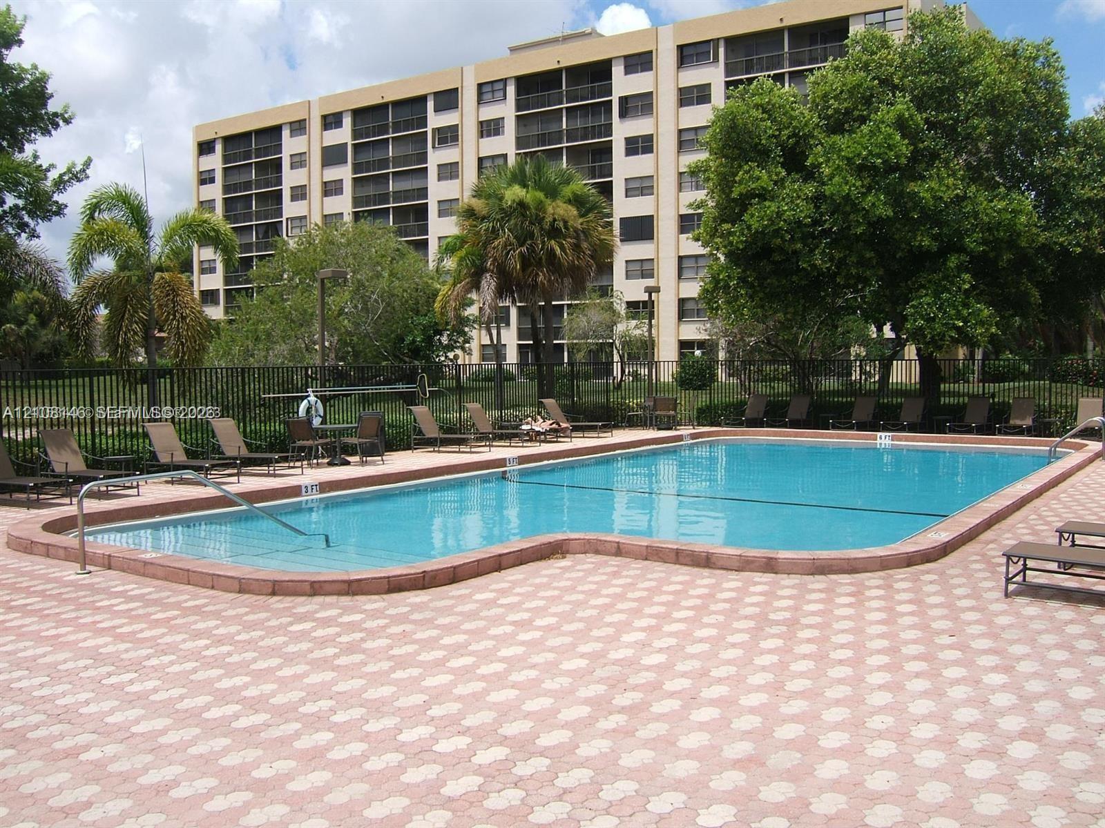 In-ground swimming pool with a curved edge and integrated metal handrail, surrounded by a geometric-patterned stone paver deck, mature landscaping, and perimeter metal fencing