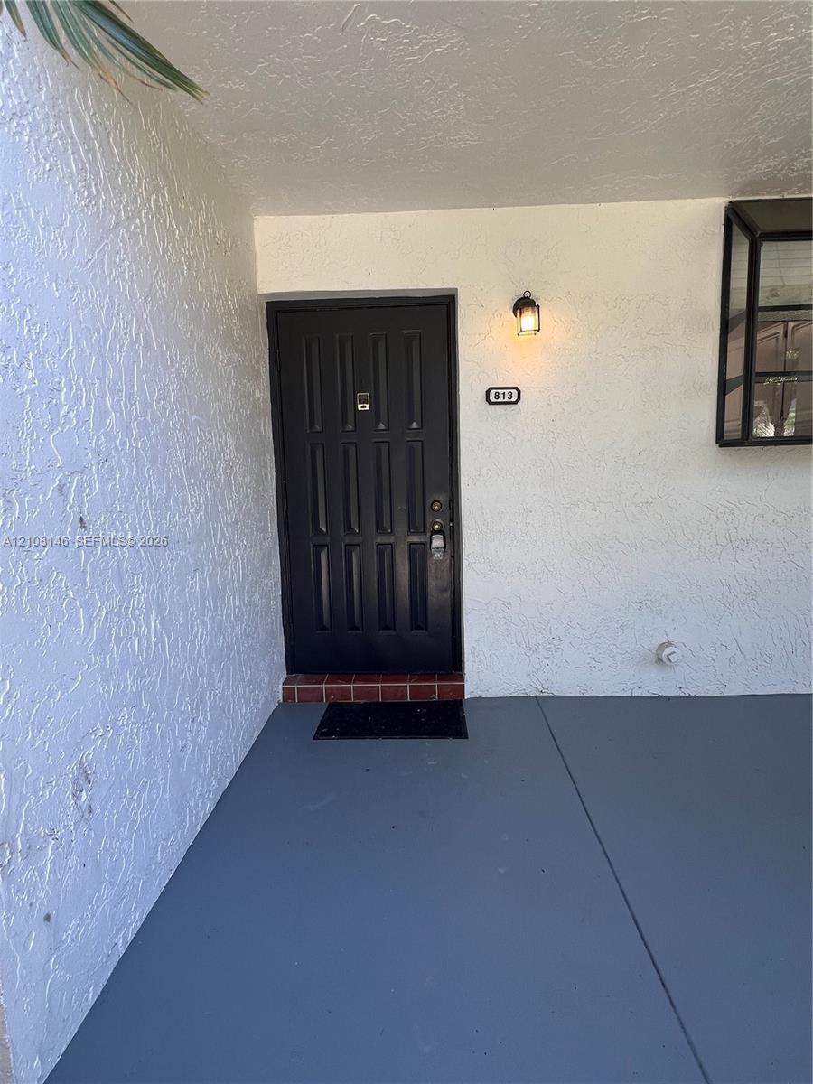 Covered entryway featuring a textured stucco facade, dark-toned paneled front door, brick threshold, and wall-mounted exterior lighting