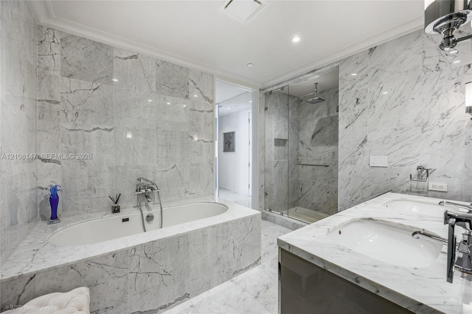 Bathroom featuring floor-to-ceiling stone tile surround, soaking tub with wall-mounted hardware, glass-enclosed walk-in shower, and dual-basin vanity with stone countertops