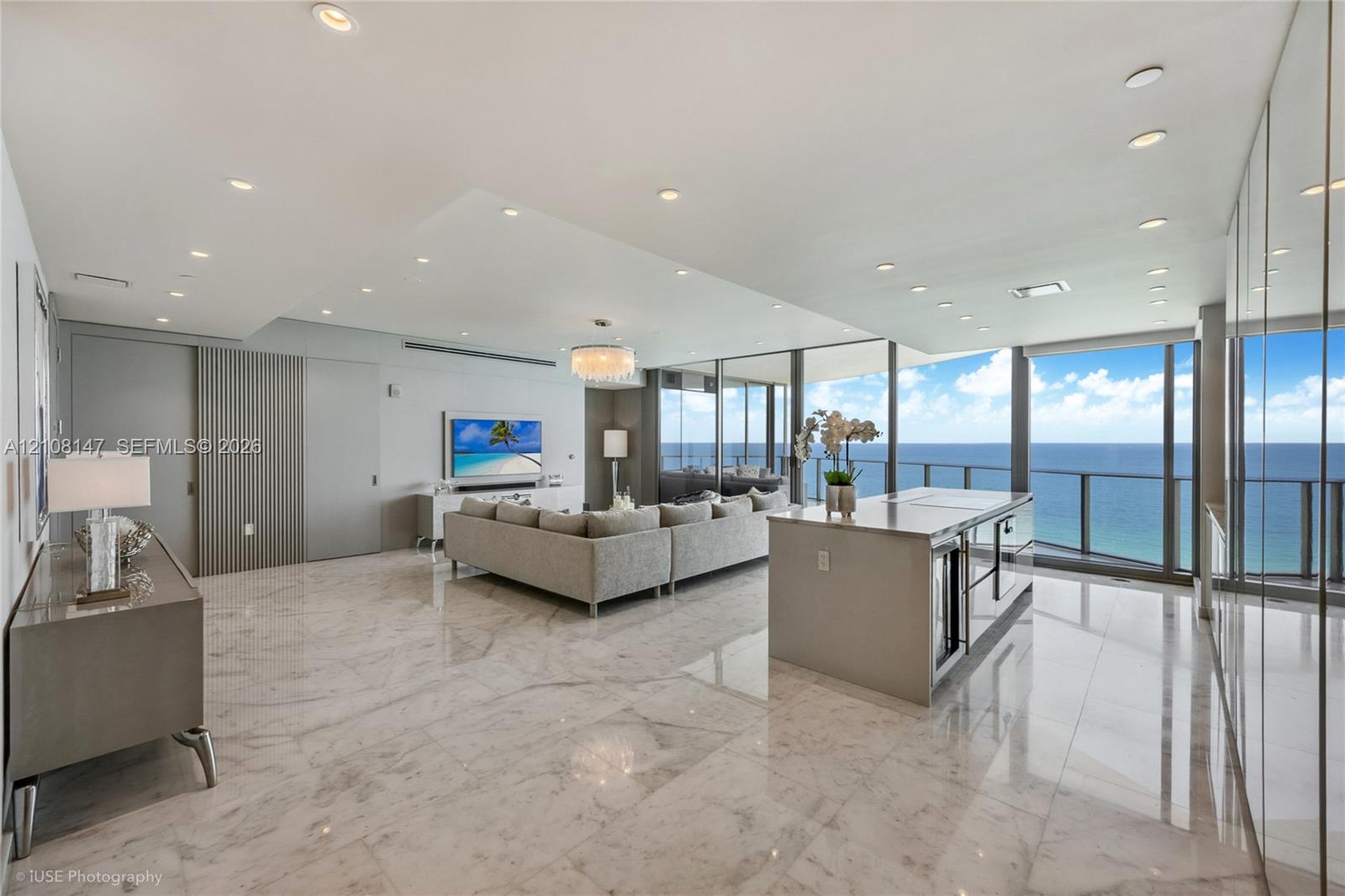 Open-concept living space featuring polished stone flooring, floor-to-ceiling glass wall panels, an integrated kitchen island with stone countertop, recessed ceiling lighting, and vertical-slat wall paneling