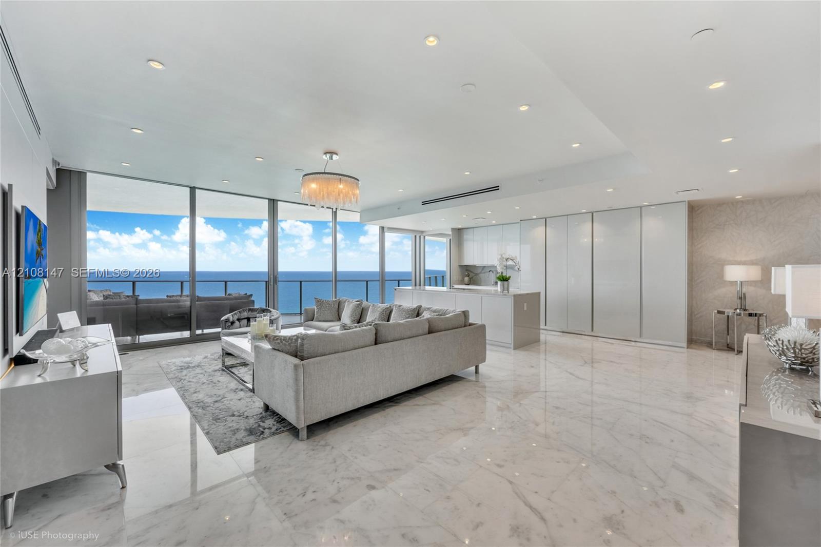 Open-concept interior featuring floor-to-ceiling glass panel walls, light-toned stone-finish flooring, recessed lighting, sleek high-gloss cabinetry, and an integrated kitchen island