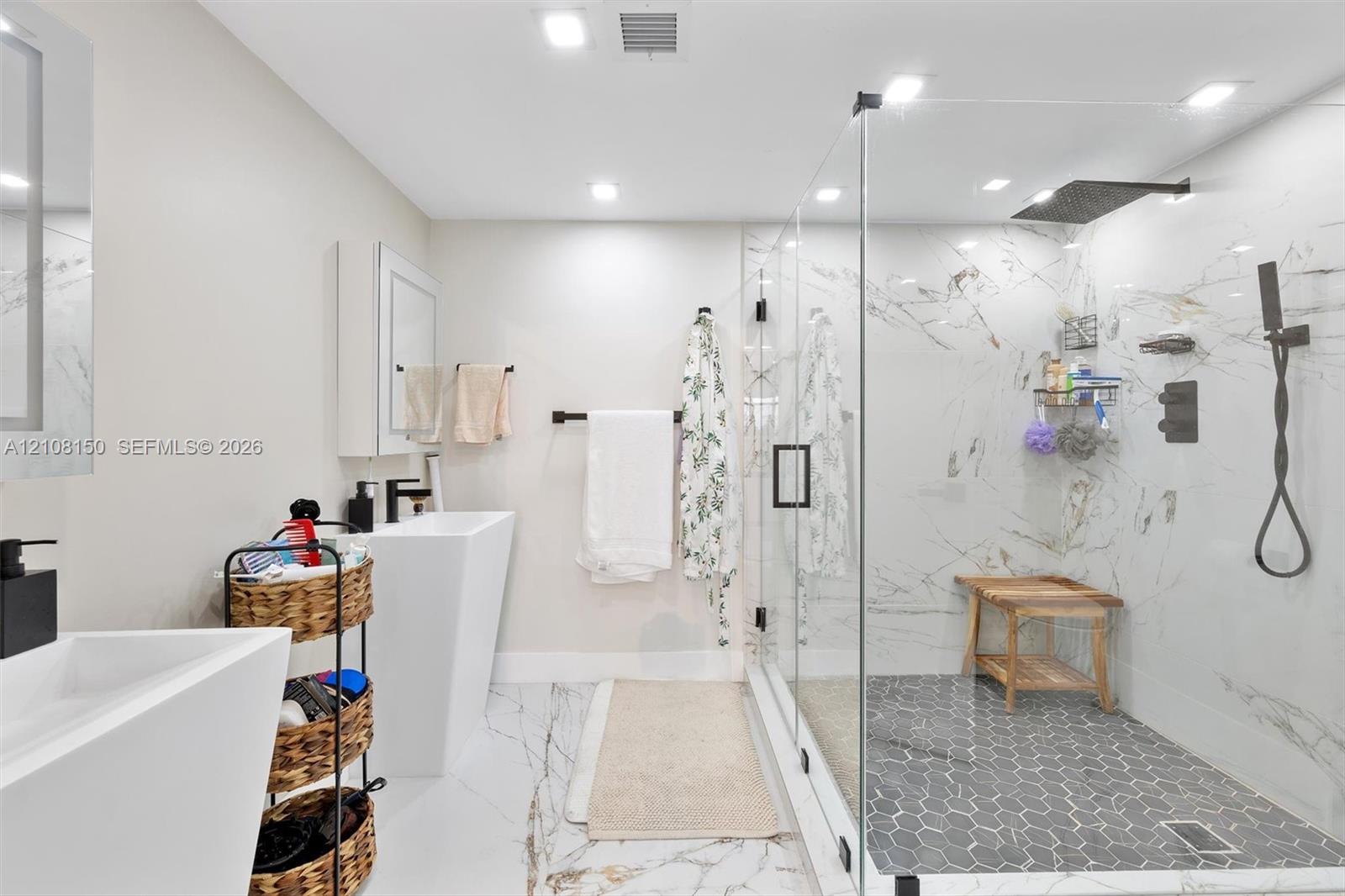 Bathroom featuring large-format stone-finish flooring, pedestal sinks, and a walk-in glass-enclosed shower with contrasting hexagon tile flooring, stone-finish wall cladding, and matte black rainfall showerhead