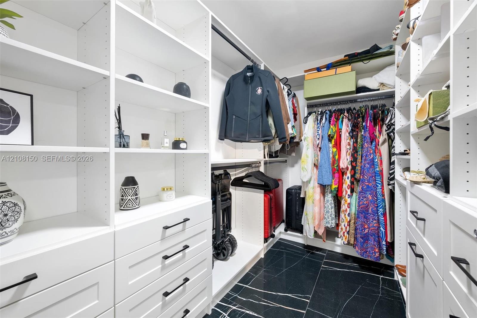Custom walk-in closet featuring white built-in shelving, floor-to-ceiling storage units, multiple drawers with matte black hardware, integrated hanging rods, and dark stone-finish tile flooring