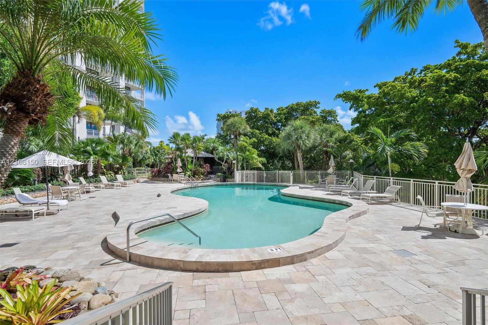 Free-form swimming pool with stone-tiled decking, metal safety fencing, and surrounding mature palm trees and tropical landscaping