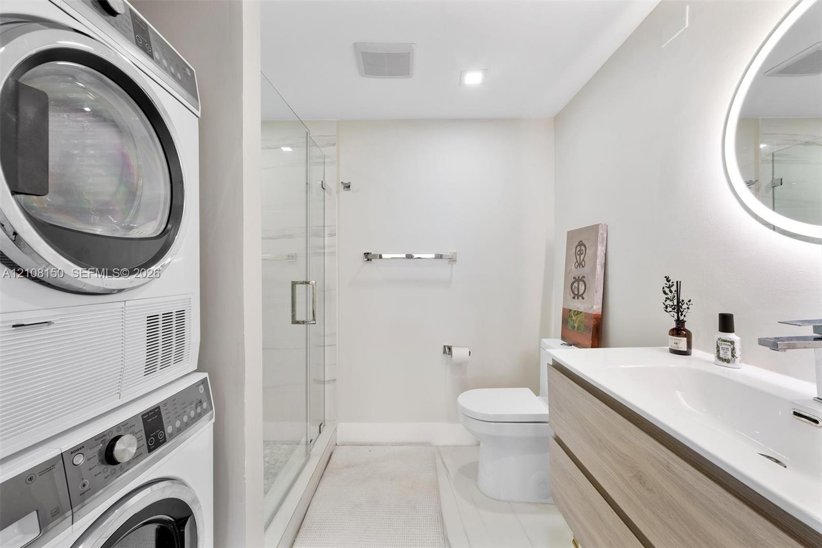 Stacked laundry units, walk-in shower with frameless glass enclosure, wall-mounted floating vanity with integrated sink, and illuminated vanity mirror