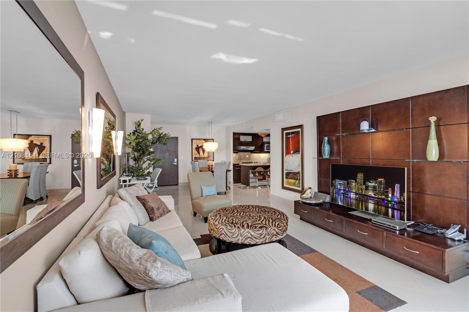 Open-concept living space featuring a floor-to-ceiling built-in dark wood media wall with integrated shelving, large wall-mounted mirror, neutral tile flooring, and wall-mounted sconce lighting