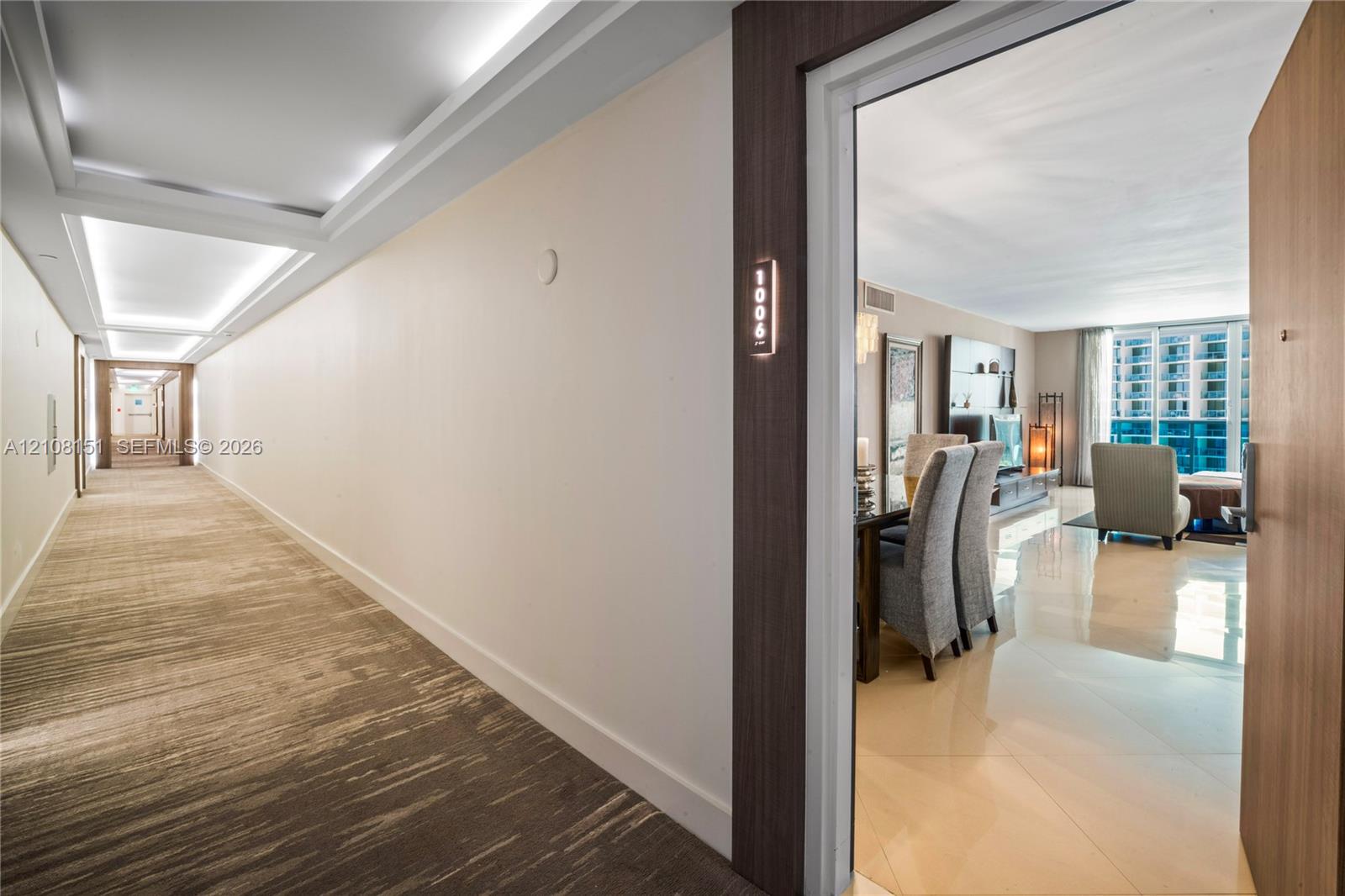 Interior hallway featuring recessed ceiling lighting and patterned carpeting, opening to an entryway with light-colored tile flooring and a wall-mounted unit number plate