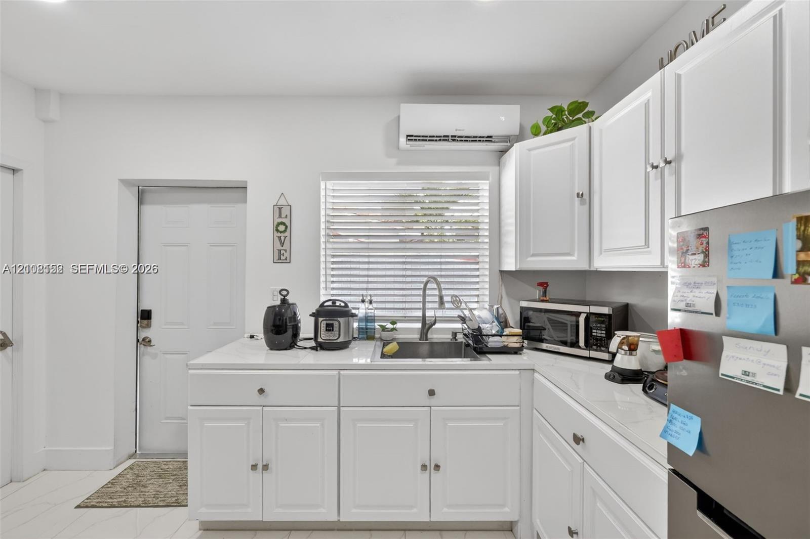 Kitchen area featuring white shaker-style cabinetry, solid-surface countertops, an undermount stainless steel sink with a pull-down faucet, and a wall-mounted climate control unit