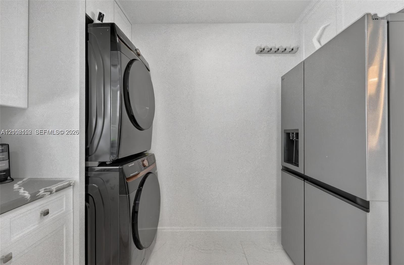 Utility space featuring white cabinetry, a neutral stone-finish countertop, stainless steel refrigerator, and stacked laundry appliances