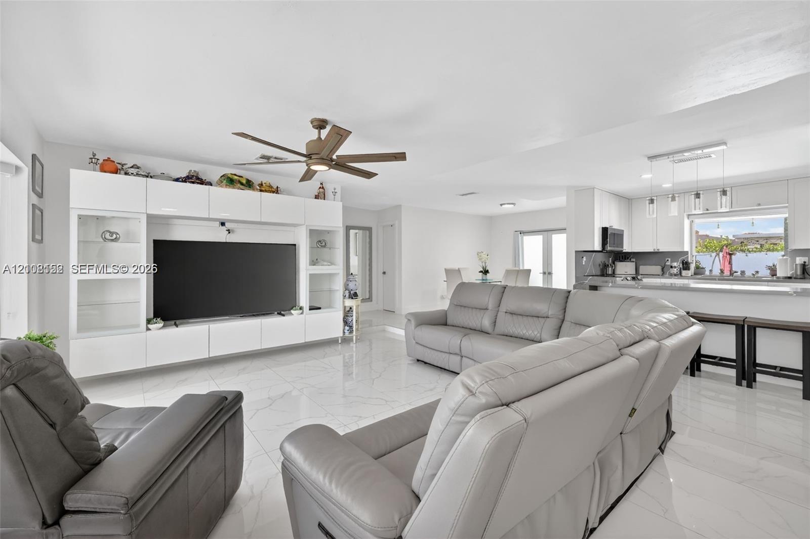 Open-concept interior featuring large-format polished tile flooring, white built-in media cabinetry, a modern ceiling fan, and a breakfast bar