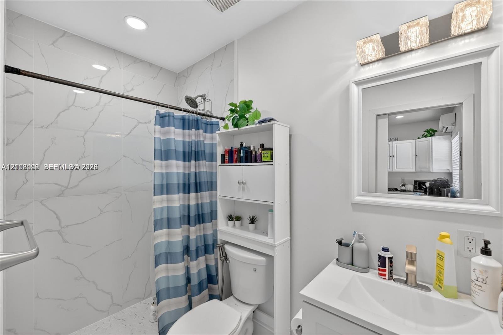Full-height wall tiling with a white stone-pattern finish, recessed ceiling lighting, three-light decorative wall sconce, framed vanity mirror, and single-basin vanity with a metallic faucet