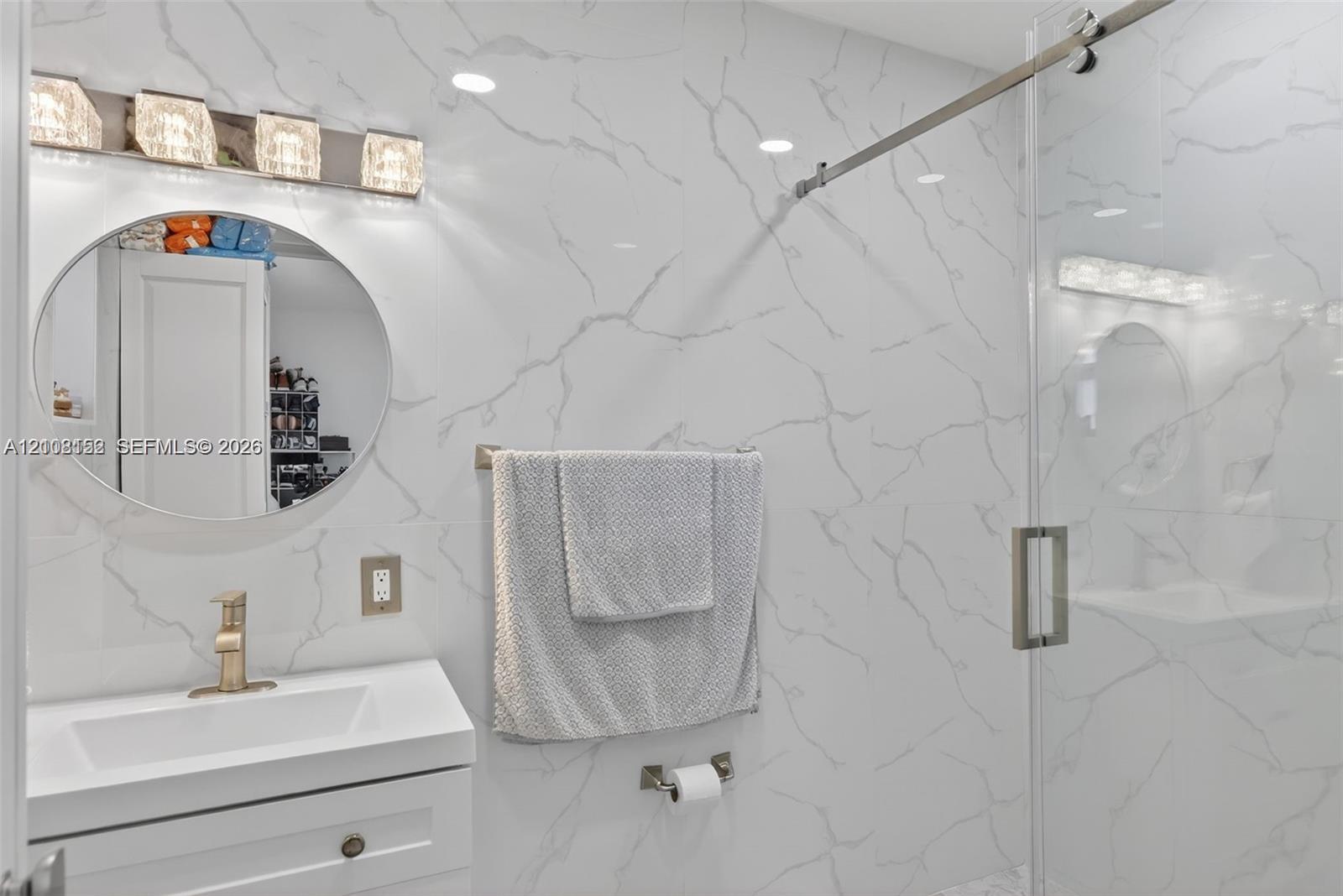 Bathroom featuring large-format wall tiling with veining patterns, a vanity with an integrated rectangular sink, a circular mirror, a glass-enclosed shower with metal hardware, and a four-light fixture