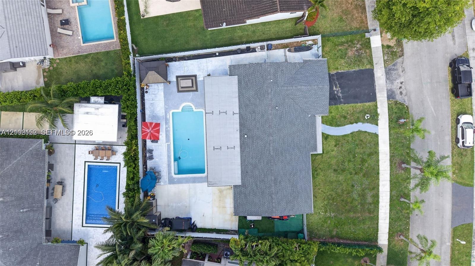 Aerial view featuring a rectangular swimming pool, concrete patio, asphalt driveway, and landscaped front lawn