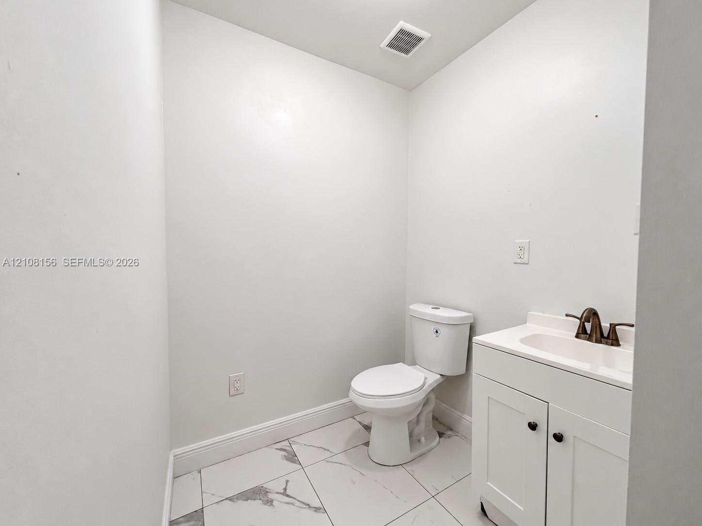 White vanity with a neutral countertop, dark metal faucet, ceramic toilet, and large-format tile flooring with a veined pattern