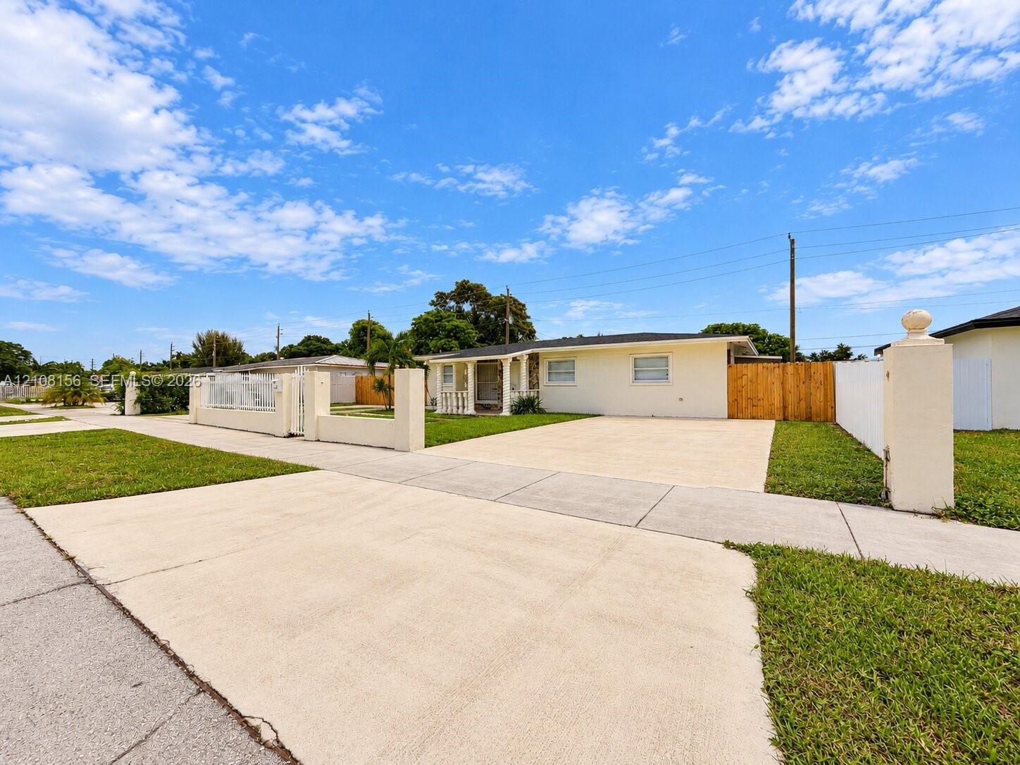 Single-story exterior featuring a light-colored stucco facade, expansive concrete driveway, gated front perimeter wall, and wood-panel privacy fencing