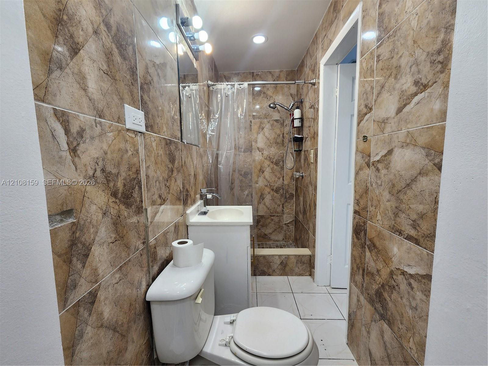 Full-height wall tiling with a decorative stone pattern, white vanity with an integrated sink, walk-in shower with a handheld spray head, and recessed ceiling lighting