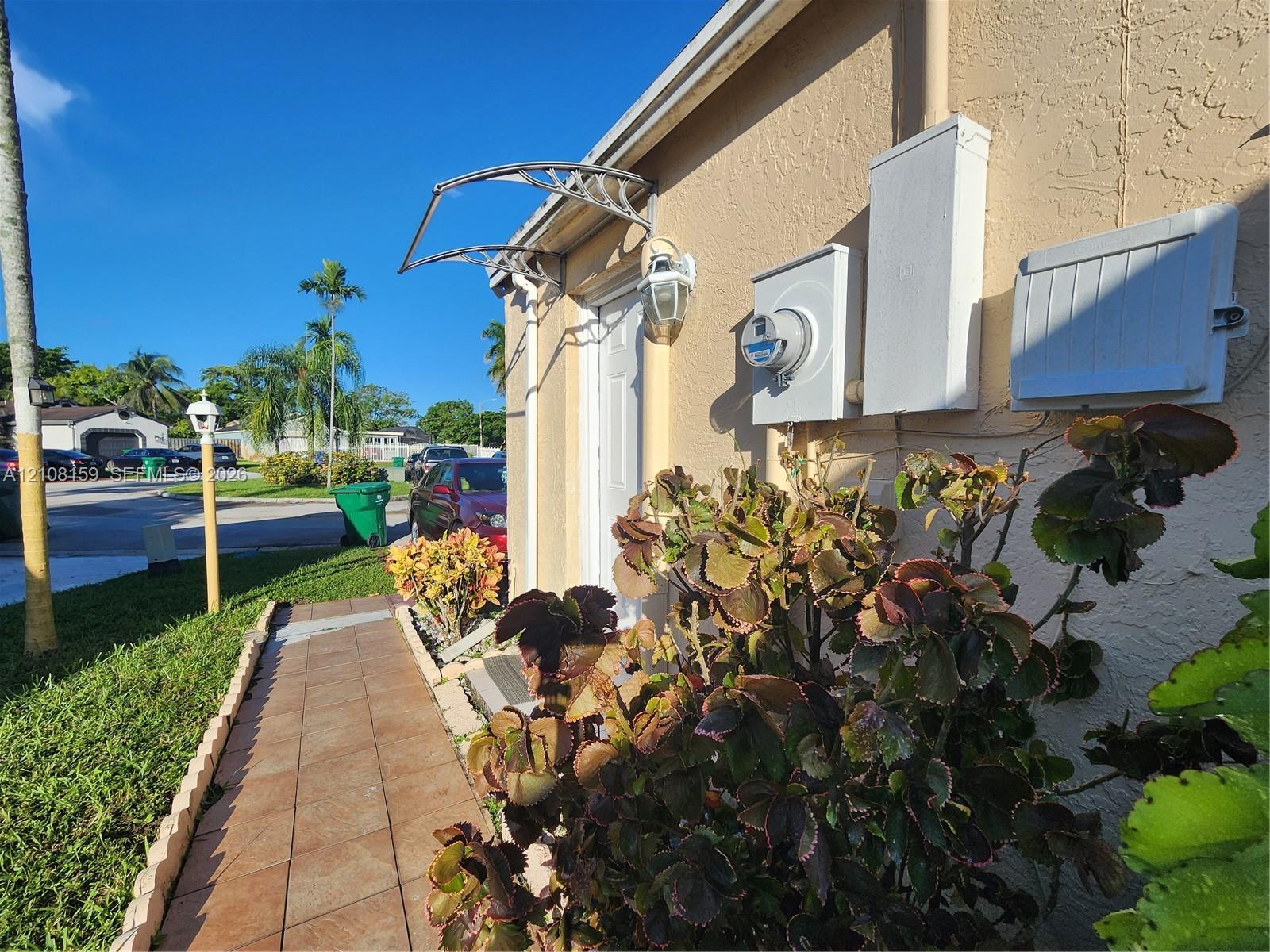 Stucco exterior featuring a tiled walkway, metal door awning, exterior wall-mounted light fixture, and mature landscape plantings