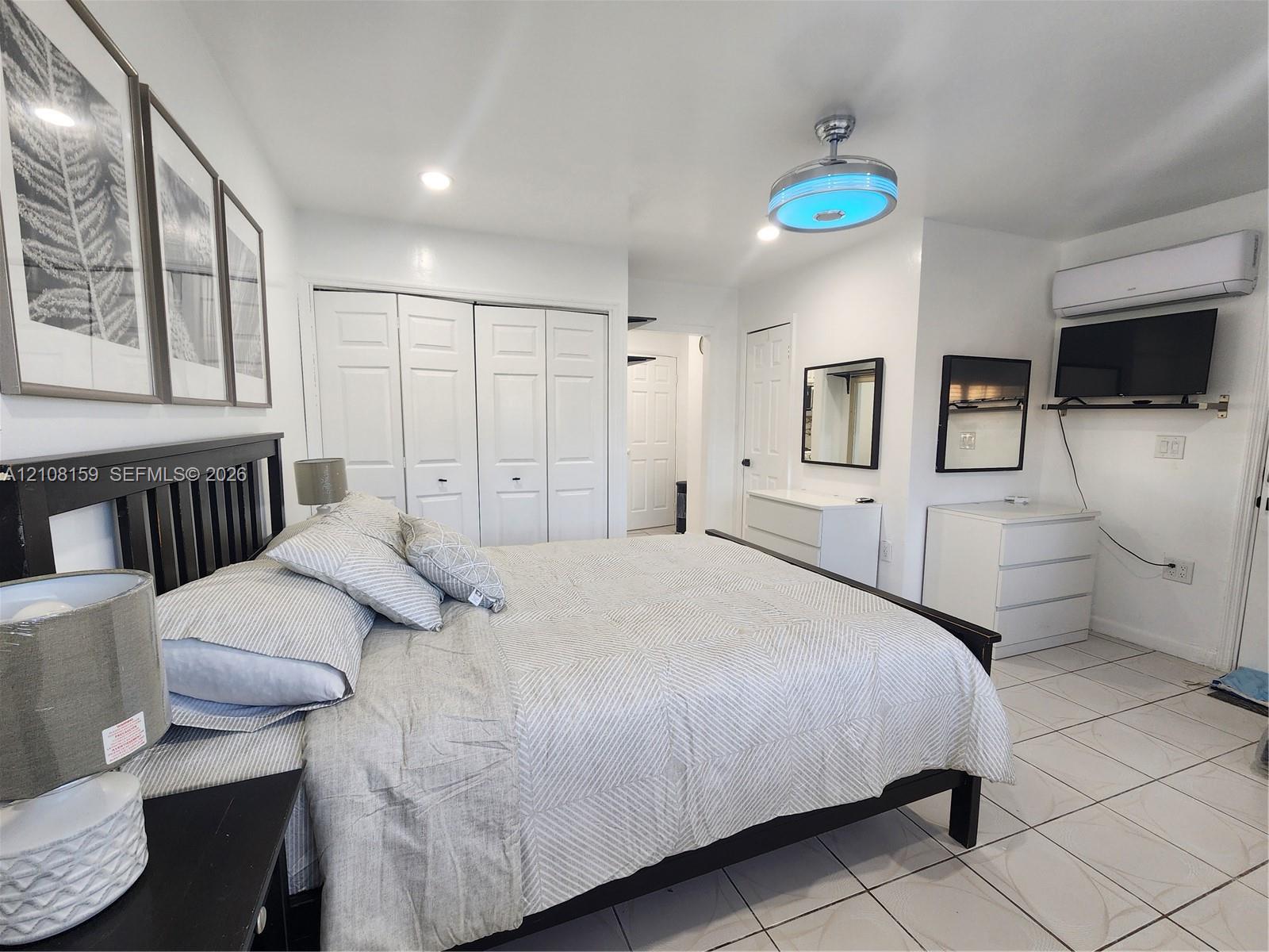 Neutral interior featuring white tile flooring, multi-panel sliding closet doors, ceiling-mounted light fixture with fan, and a wall-mounted ductless climate control unit