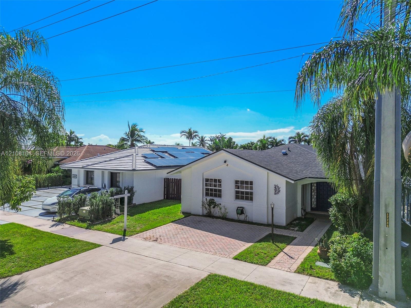 Single-story exterior featuring white stucco siding, a shingled roof with integrated solar panels, paved brick driveway, and mature landscaping