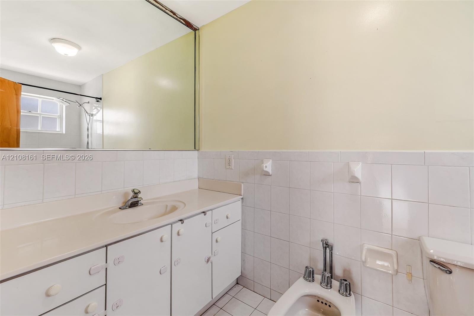 Bathroom featuring a white vanity with an integrated sink, a full-width wall mirror, white wall tiling, and a bidet